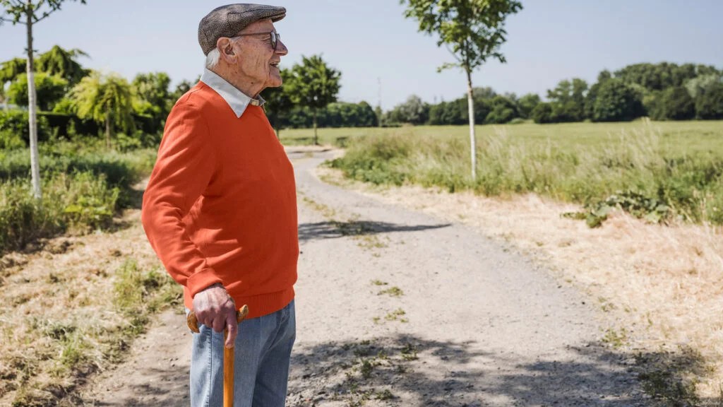 Un homme plus âgé portant un chapeau et tenant une canne regarde au loin tout en se promenant dehors