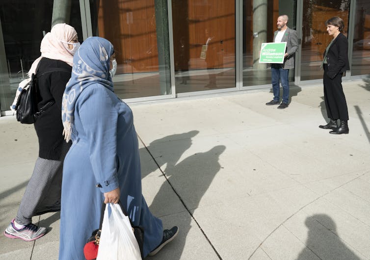 Deux femmes portant un foulard et portant des sacs de courses passent devant un homme politique.