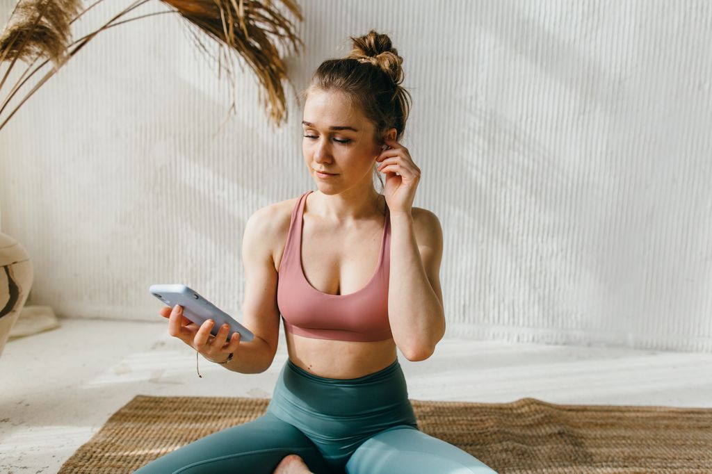 Une jeune femme portant des écouteurs sans fil utilise son smartphone pour configurer une application de méditation, configure une liste de lecture musicale avant une séance d'entraînement et sélectionne un programme de remise en forme approprié en ligne