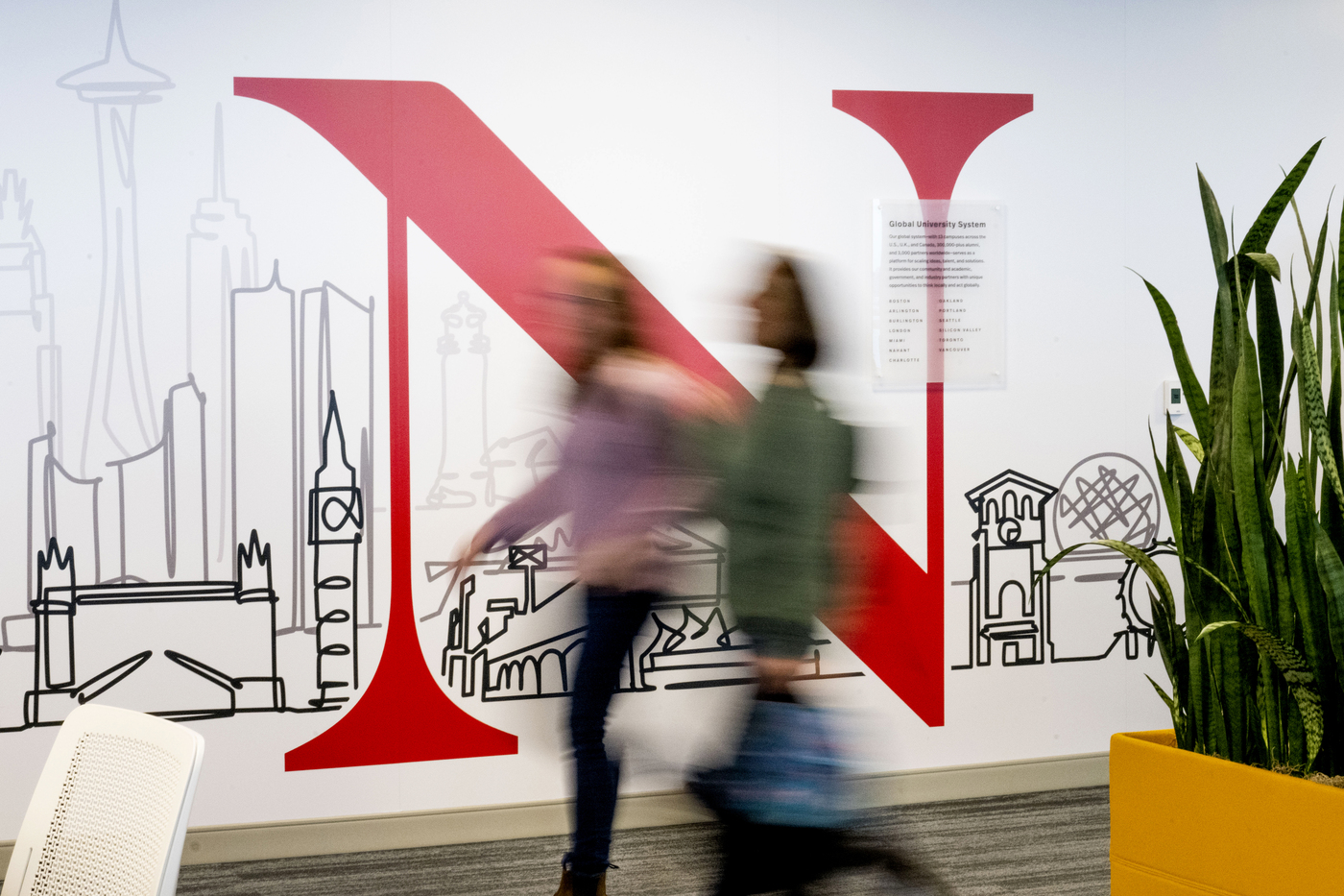 A time lapse photo of two people walking in front of a Northeastern logo at the Portland campus.
