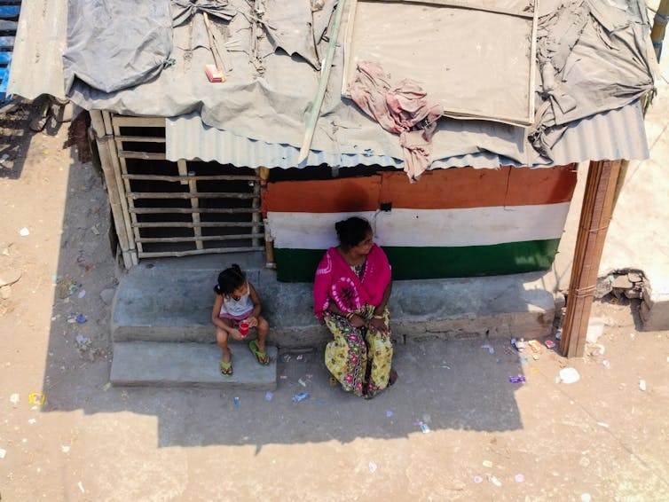 Une femme et une jeune fille sont assises à l’ombre devant un petit bâtiment aux portes à lattes et au toit précaire.