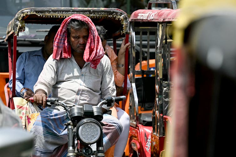 Un homme plus âgé conduisant un pousse-pousse électrique avec deux passagers est assis en plein soleil dans la circulation par une chaude journée.  Il avait un bandana sur la tête pour se protéger.