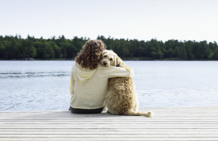 Une femme est assise sur un quai, les bras autour de son chien, qui regarde par-dessus son épaule vers la caméra.