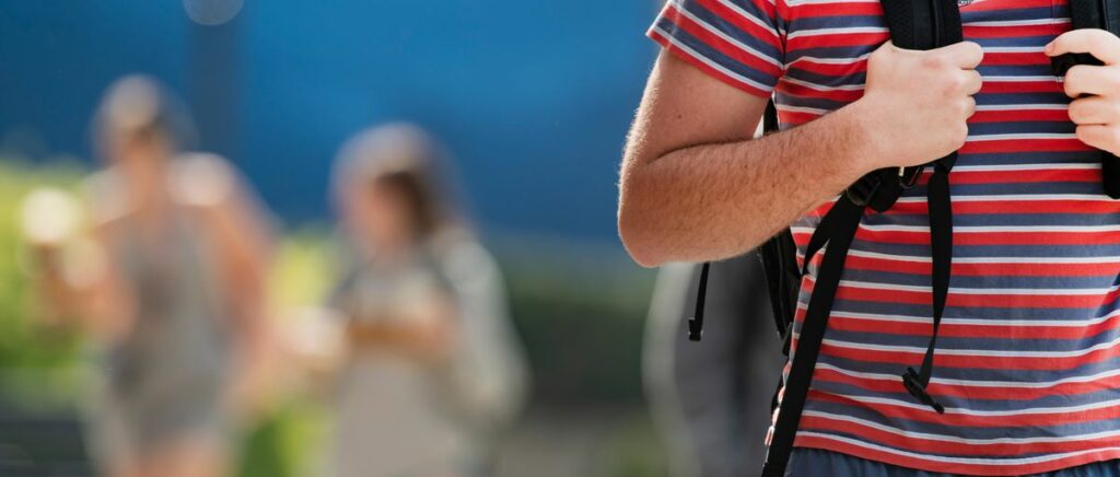 A male student is shown tightly cropped in the foreground. His head is out of the frame but he is wearing a red and blue striped shirt, blue shorts, and a gray and black backpack. His hands are holding the straps on his chest. There are two other people