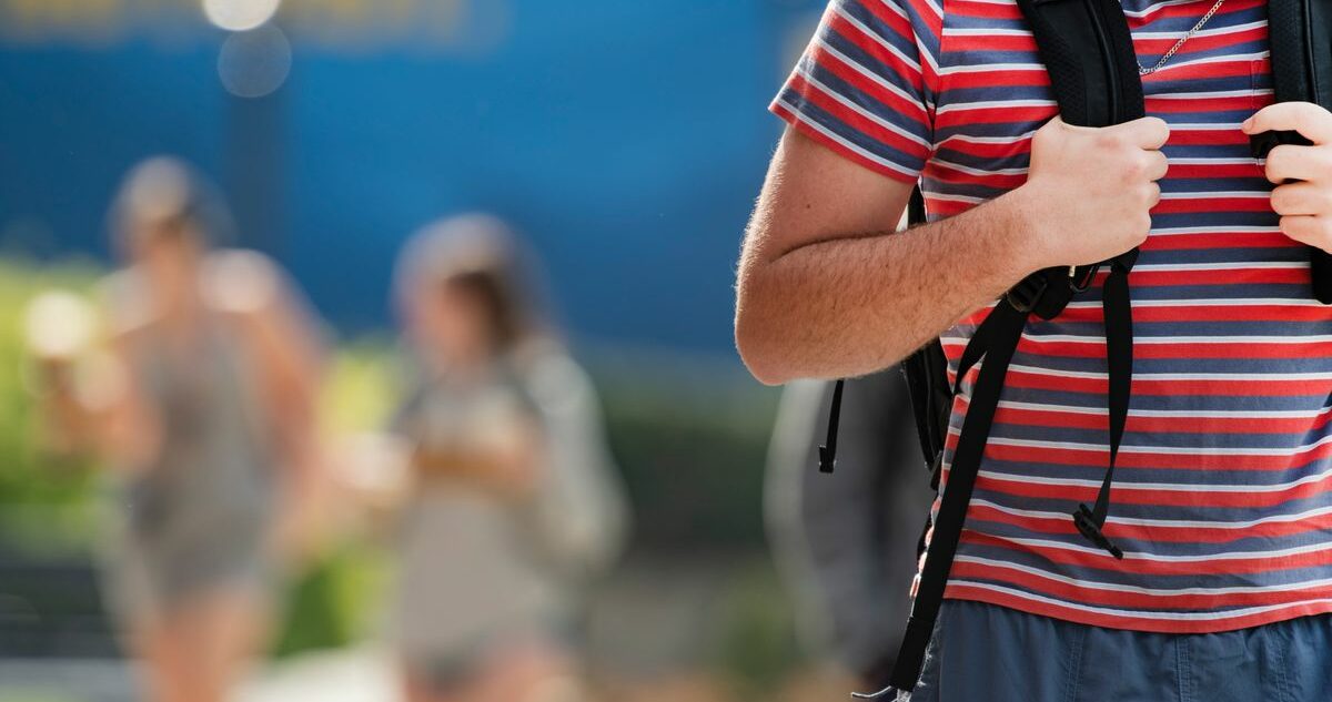 A male student is shown tightly cropped in the foreground. His head is out of the frame but he is wearing a red and blue striped shirt, blue shorts, and a gray and black backpack. His hands are holding the straps on his chest. There are two other people